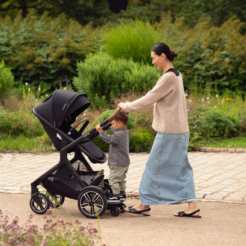 Mom pushes toddler on rider board of Nuna DEMI Next Stroller and PIPA Aire RX Car Seat Travel System - Caviar