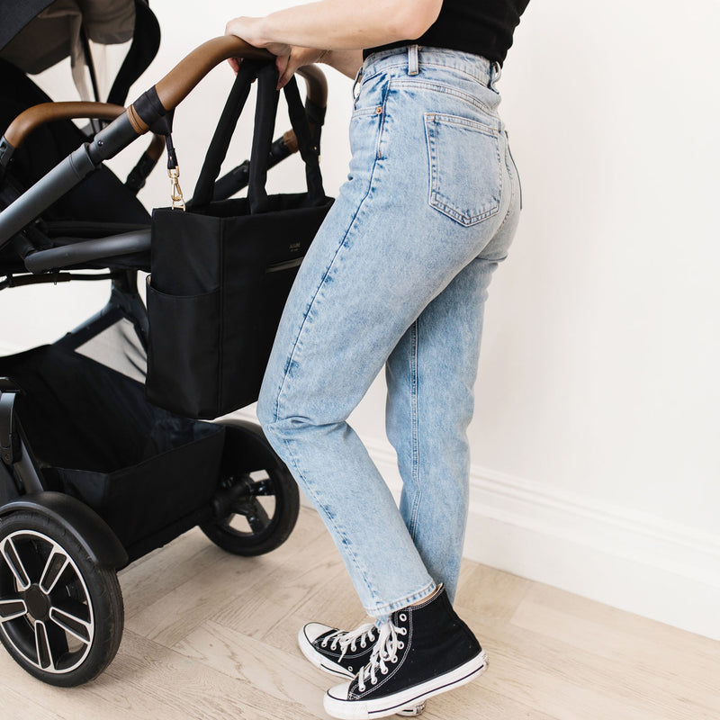 Woman pushes stroller with JuJuBe Utility-Strap Tote - Black attached to handlebar