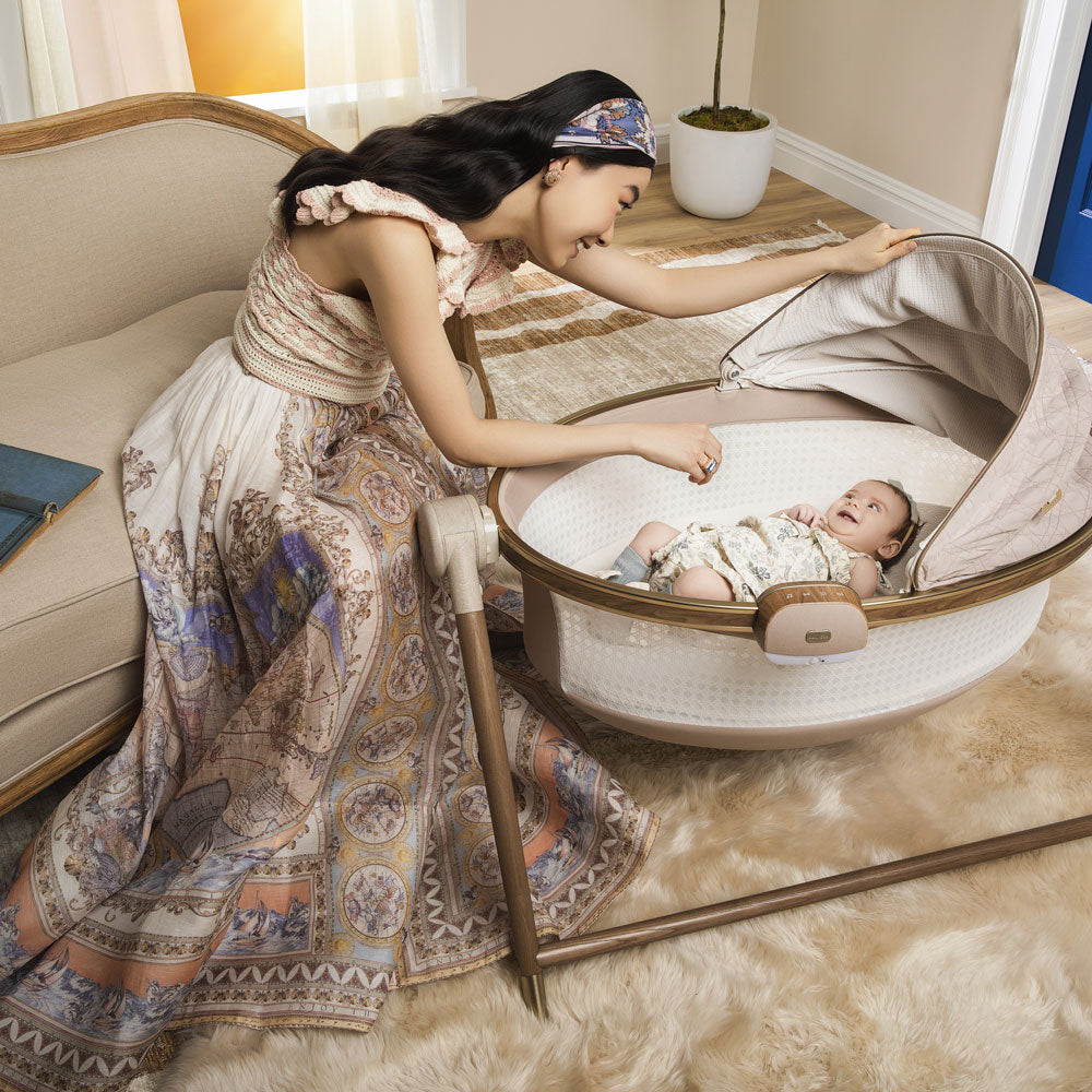 Woman smiles at baby lying in Maxi-Cosi Sibia Bassinet - Natural Heritage