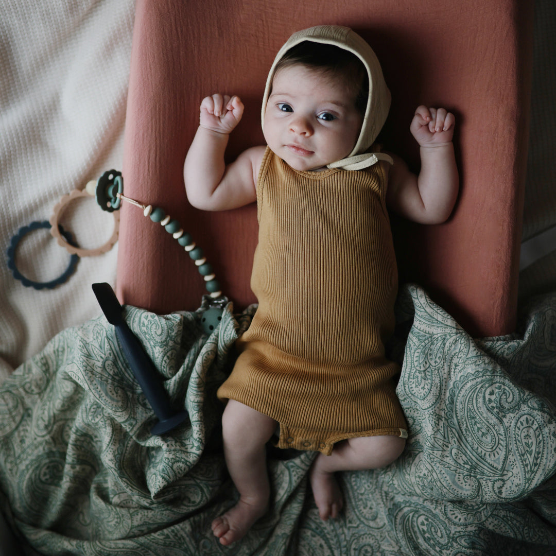 Baby lying next to Mushie Silicone Pacifier Clip - Luna - Dried Thyme