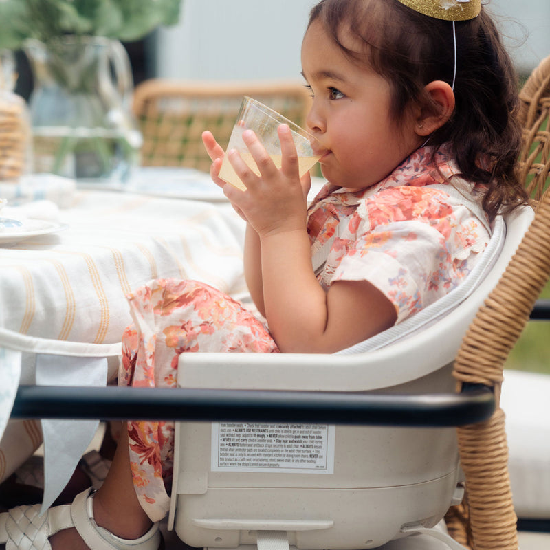 Toddler girl at party sits in Maxi-Cosi Moa 8-in-1 High Chair - Classic Oat while drinking lemonade