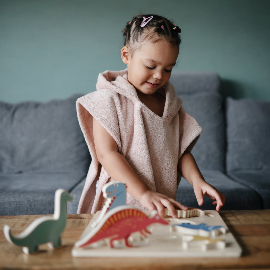 Little girl plays with dinosaur puzzle while wearing Mushie Bear Poncho Towel - Blush