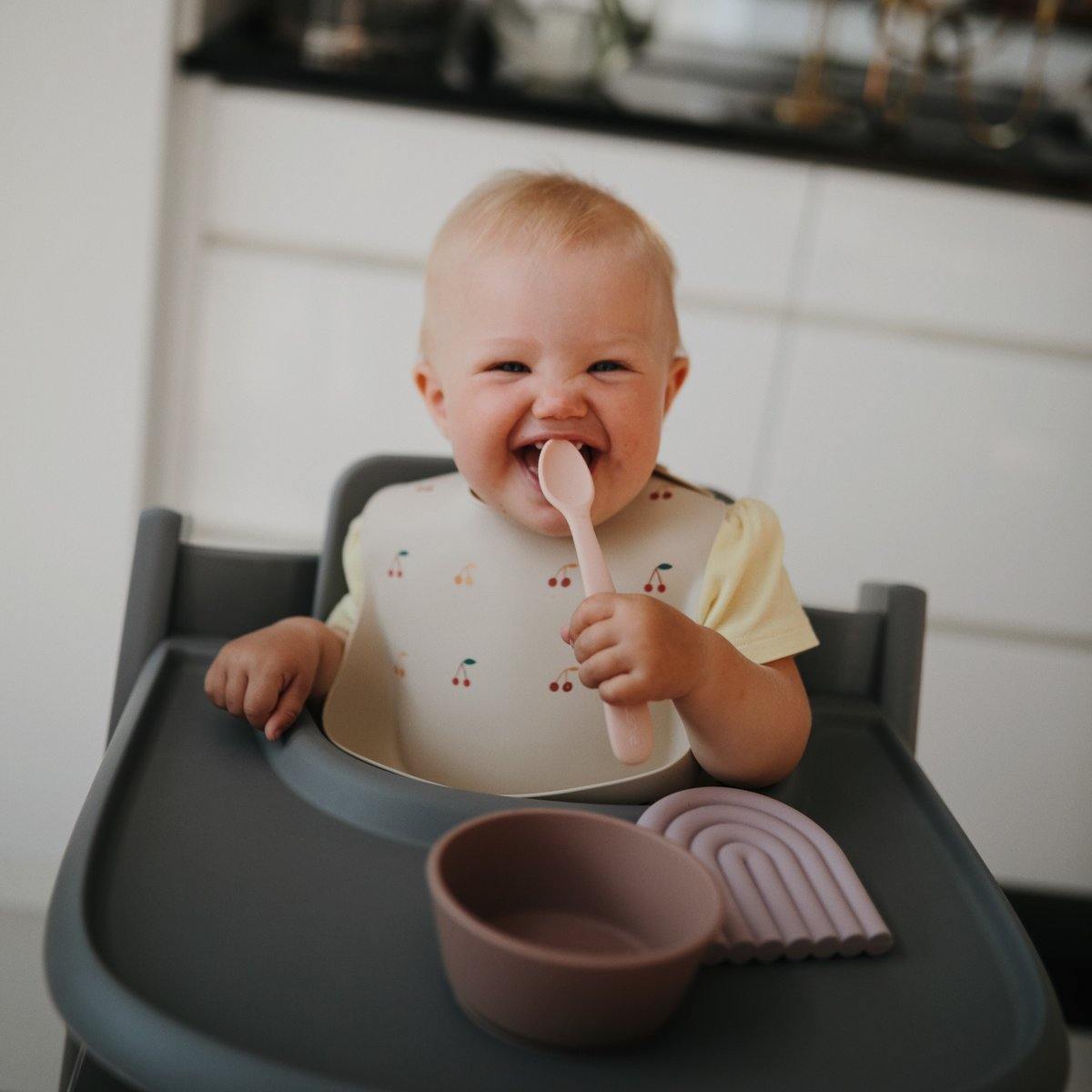 Baby smiling and chewing on Mushie Silicone Feeding Spoons - 2 Pack